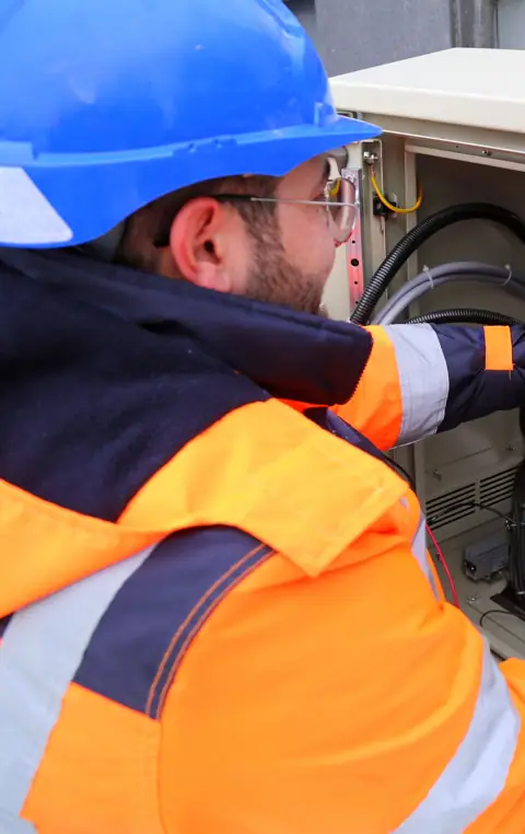 Electrician in orange hi-vis and a blue hard hat working at an outdoor electrical board
